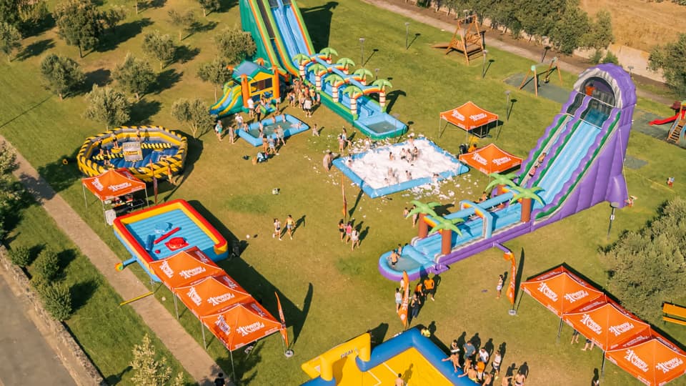 Vista dall'alto di un Acqua Party Happy Island con scivoli ad acqua, gonfiabili, gazebo arancioni e bambini in piazza